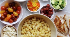 Separate ingredients in white bowls for a DIY pasta salad, including pasta, cherry tomatoes, pepper rings, turkey slices, among others.