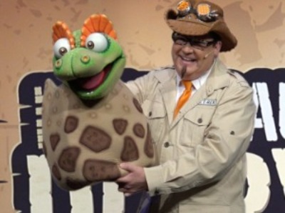 A man dressed in khaki safari clothing holds a green and orange baby dinosaur puppet that is emerging from a brown egg.