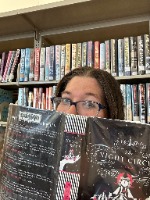A woman with brown hair and glasses sits in front of full book shelves covering the bottom half of her face with an open book.