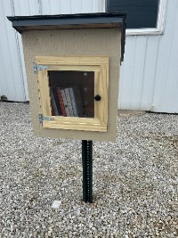 The Free Library, consisting of a wood box on a fence post, with a black roof and a windowed door.