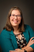 A brunette woman with glasses wears a black and white top with a teal jacket and poses for a professional headshot.