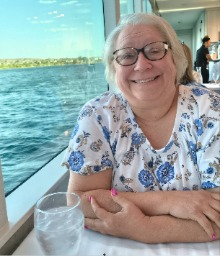 A blond woman wearing glasses and a white shirt with blue flowers sits near a window on a boat and smiles at the camera.